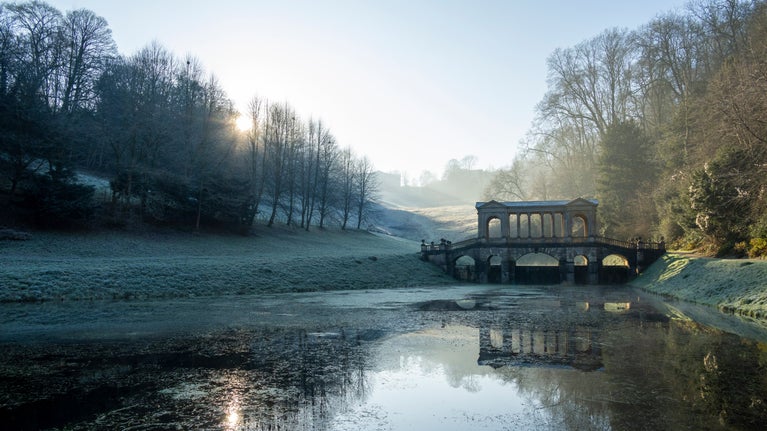 A wintery scene with frosty grass and low sun shining behind the Palladian Bridge at Prior Park Landscape Garden, Somerset